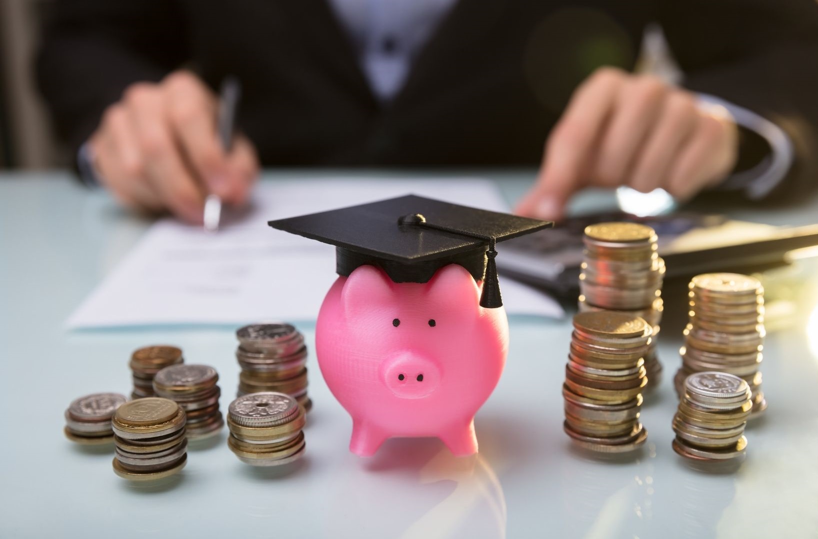 small piggy bank with a graduation cap on in the centre of the image. Money coins are stacked either side and in the background is someone studying